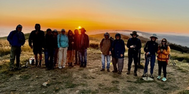 A group of hikers stand at the top of Borel Hill in Russian Ridge Open Space Preserve with the sun setting behind them