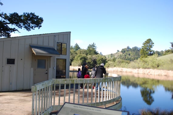 Children and parents stand on the deck of the Daniels Nature Center overlooking Alpine Pond